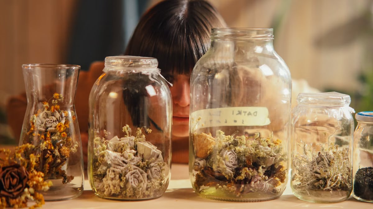 A woman thoughtfully looks at dried flowers arranged in multiple glass containers on a table.