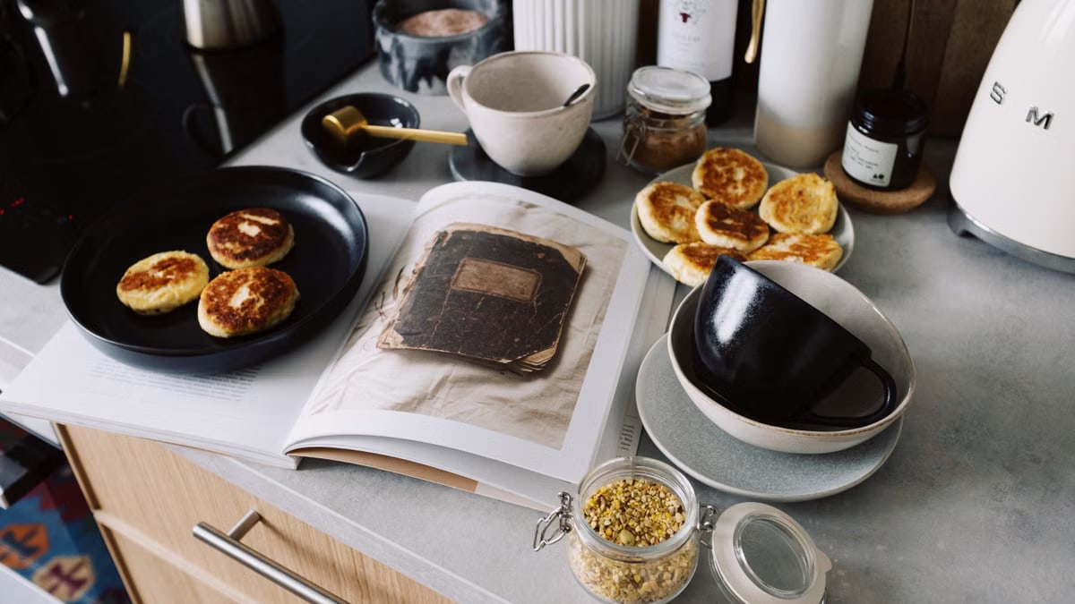 High angle of cookbook and cooked fritters on plates placed on table near utensils and ingredients in cozy kitchen at home